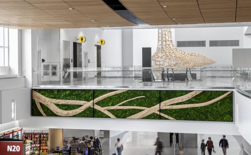 “Airport concourse with travelers below a large green wall art installation featuring tan, branch‑like patterns. Above it is a glass‑railed level with restrooms and a large sculptural structure made of light-colored latticework.