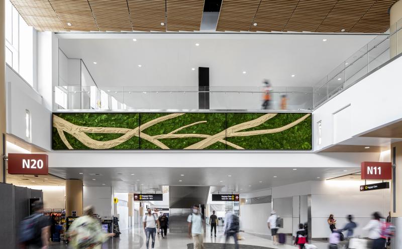 Airport concourse with travelers walking below a large green wall art installation featuring tan, branch‑like patterns. Overhead is a glass‑railed walkway, and signs for gates N20 and N11 are visible.