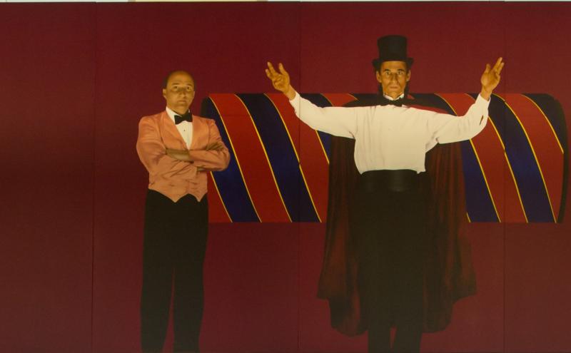 Painted panel showing the magician with arms raised before a large rolled curtain, the assistant standing with arms crossed beside him.  