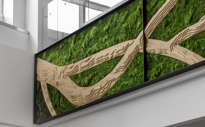 Close-up of a green wall art installation featuring layered tan, branch‑like forms set against textured green foliage, with a glass‑railed upper level visible above.