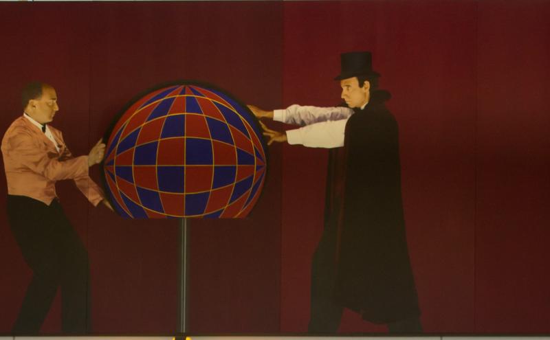 Painted panel showing magician and assistant each reaching toward a large sphere on a stand between them.
