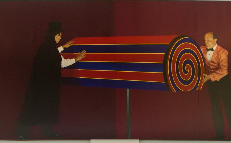 Painted panel showing the magician and assistant with a large horizontal cylinder between them, its end face showing a spiral pattern. 
