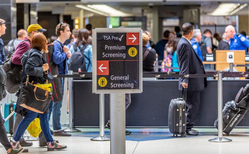 Airport security area with travelers lining up near a sign directing passengers to TSA PreCheck premium screening or general screening.
