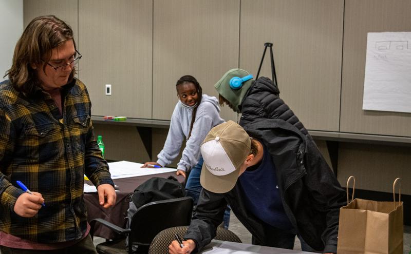 Several individuals working together at tables in a classroom or meeting room, writing on large sheets of paper.