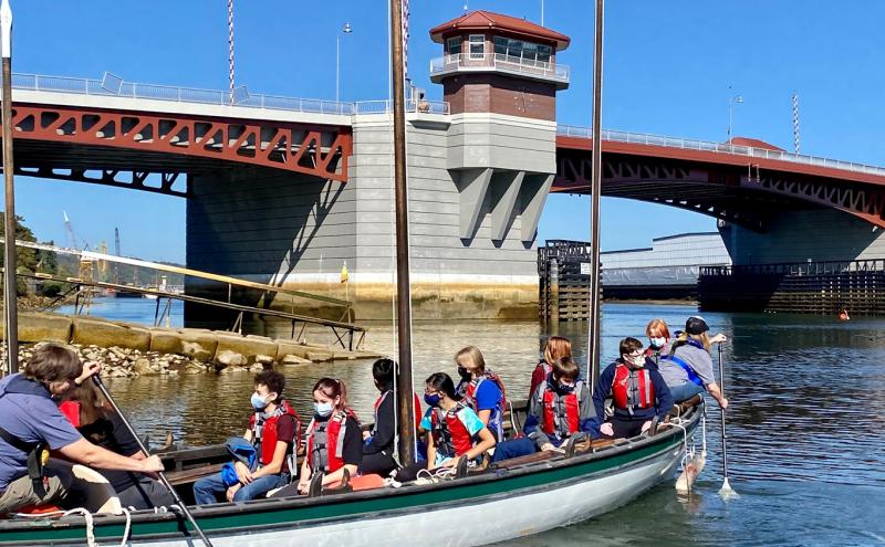 A group of individuals wearing life jackets sit in a long wooden rowing boat on calm water near a large drawbridge structure. One individual at the front and one at the back use oars to steer. The boat is close to the shoreline with rocks visible, and an industrial area with cranes appears in the background under a clear blue sky.