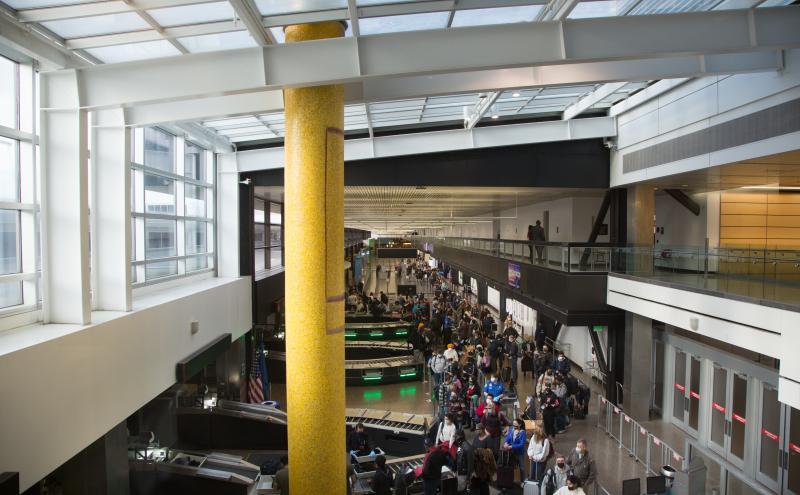 Mosaic column spanning both floors of the airport, viewed from the upper level looking down into a crowded concourse.