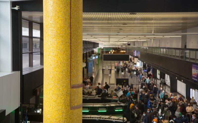 A mosaic-covered column rising the full height of the airport concourse, with a busy terminal visible behind it.