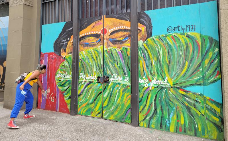 A person leans in to examine a colorful mural painted on wooden panels covering a building façade. The artwork shows a stylized face with closed eyes above sweeping green brushstrokes, with handwritten text across the center. Additional murals are visible on nearby panels along the sidewalk.