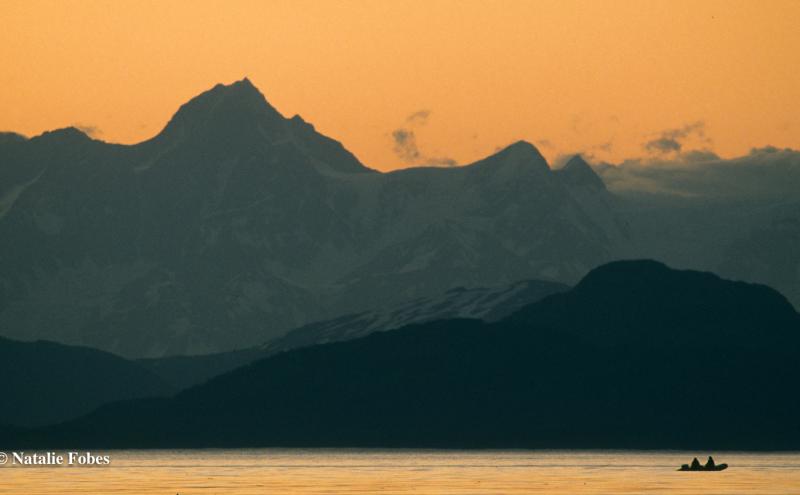 Silhouetted mountain range at sunset with an orange sky, a calm body of water in the foreground, and a small boat visible near the lower right.