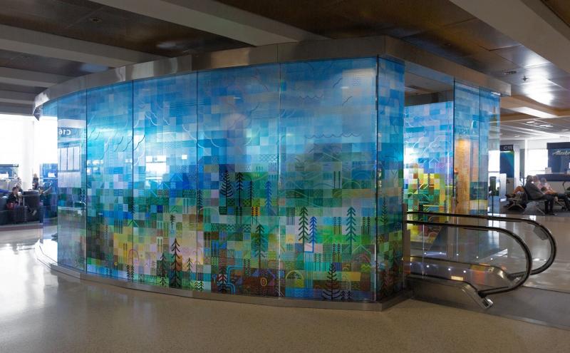 Glass panels wrapping around three sides of a escalator, forming a glowing structure in the middle of the airport concourse. 