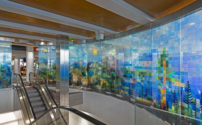 Glass panels curved around an escalator, painted with a pixelated landscape of trees and mountains. 