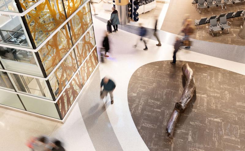 Overhead view showing the glass panel tower alongside a weather bronze log resting on the concourse floor, with travelers walking by.