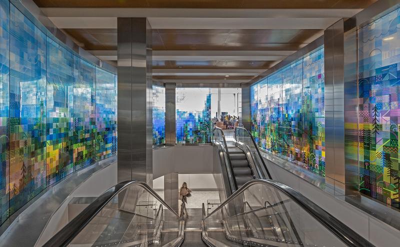 Looking down into the escalator from the entrance, with glowing painted glass panels lining both walls on either side.