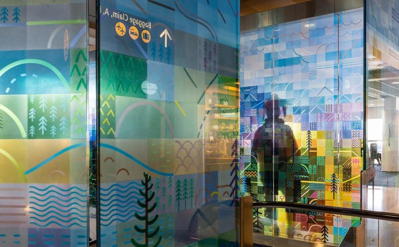 Close-up of the painted glass panels showing stylized evergreen trees, rolling hills, and wavy water lines built from flat geometric shapes, with the airport visible through the transparent glass.