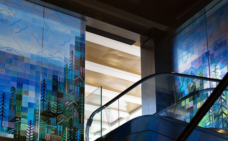 The full height of the glass panels alongside the escalator, the painted landscape running from trees and buildings at the base to the mount and clouds at the top. 