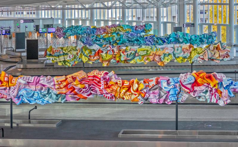 Colorful sculptural artwork made of long, ribbon‑like forms is displayed above the baggage claim area in the International Arrivals Facility.