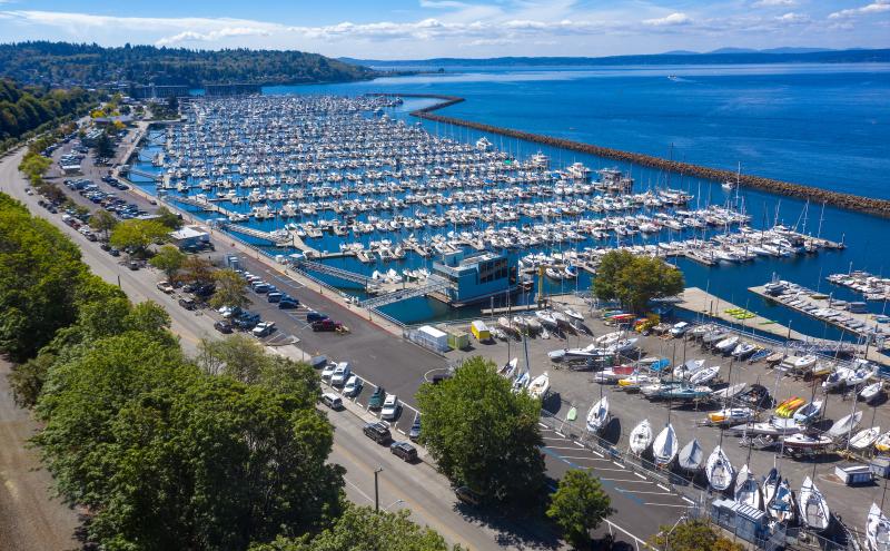 Aerial view of a large marina filled with rows of docked boats and yachts beside a long breakwater extending into a blue body of water. A parking area, trees, and additional sailboats on trailers are visible in the foreground, with forested hills and a partly cloudy sky in the background.