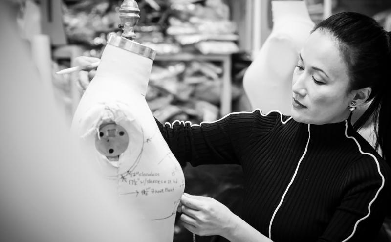 Luly Yang working in a studio, marking measurements on a dress form surrounded by sewing tools and supplies.