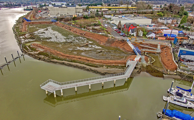 Aerial view of a waterfront restoration area with exposed shoreline, construction equipment, and a long pier extending into the water, surrounded by industrial buildings, roads, and moored boats.