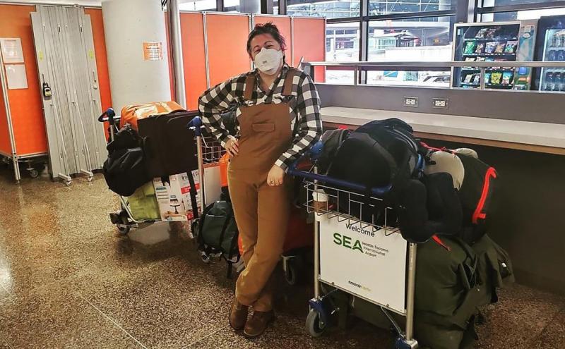 Andee Vaughan wearing a face mask stands indoors beside two airport luggage carts filled with bags and boxes. Behind the person are vending machines, windows, and orange dividers.