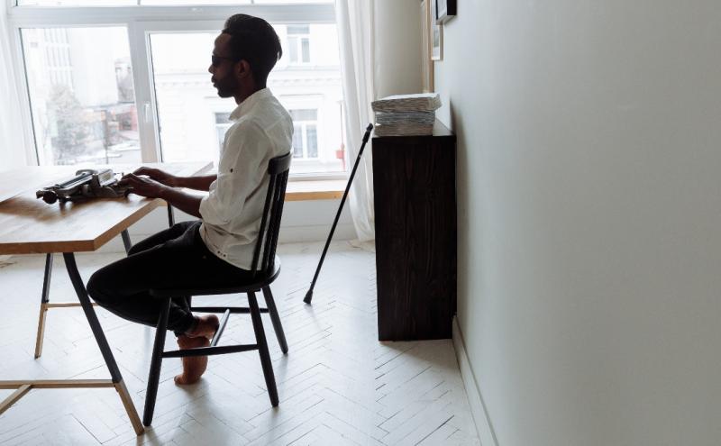 Person seated at a desk typing on a typewriter in a bright room, with a window behind the desk and a walking cane leaning against a cabinet.