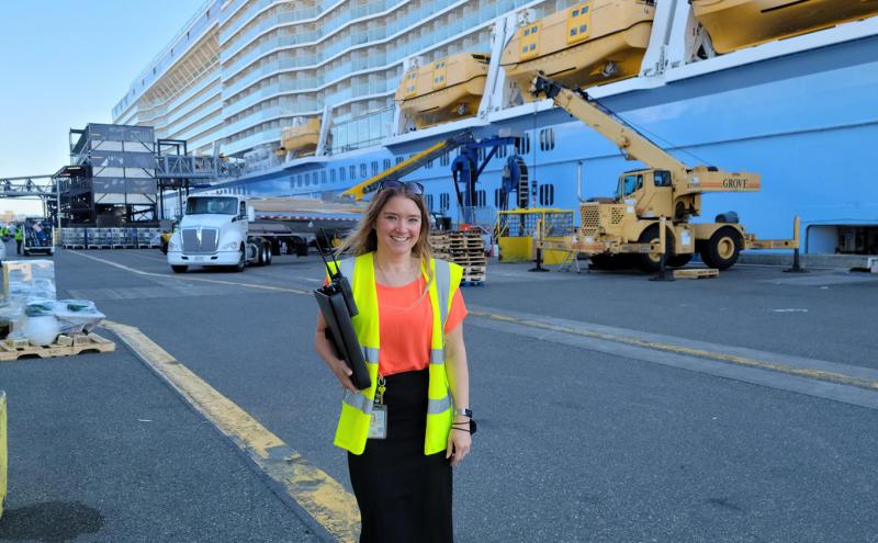 A person wearing a safety vest stands on a busy cruise ship pier with trucks, equipment, and the side of a large ship in the background.