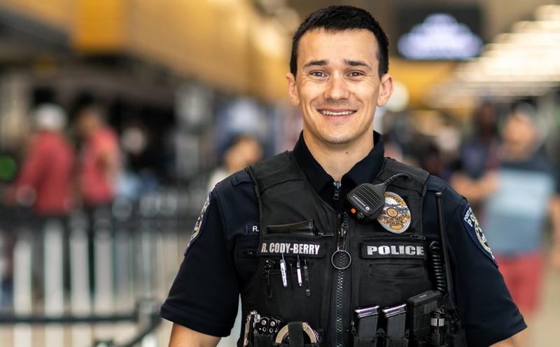 A uniformed law enforcement officer stands indoors at a busy transportation terminal, with people and blurred signs in the background.