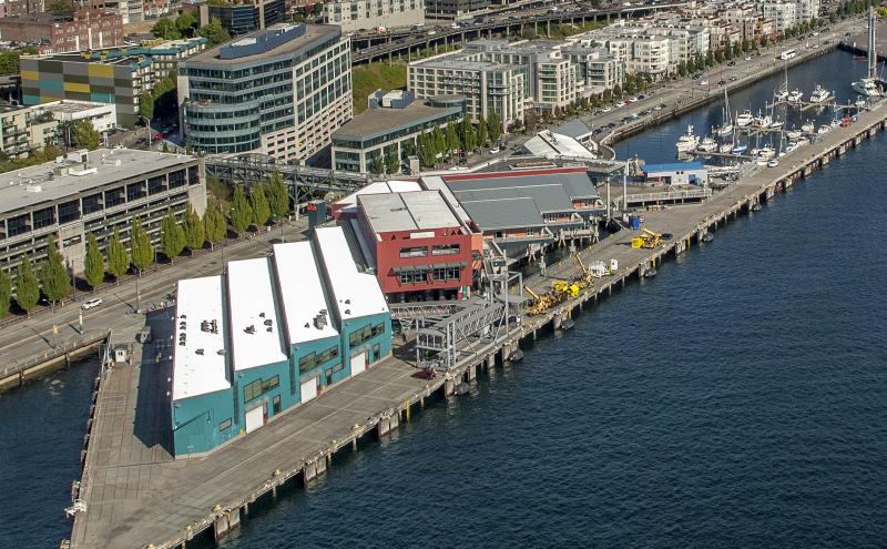 An aerial view of a waterfront pier with industrial buildings, nearby streets, and a marina with boats along the shoreline.