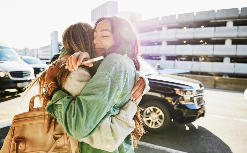 Two people embracing on an airport curbside area, with vehicles and a multi‑level parking structure in the background. One person is holding a mobile phone while carrying a backpack.