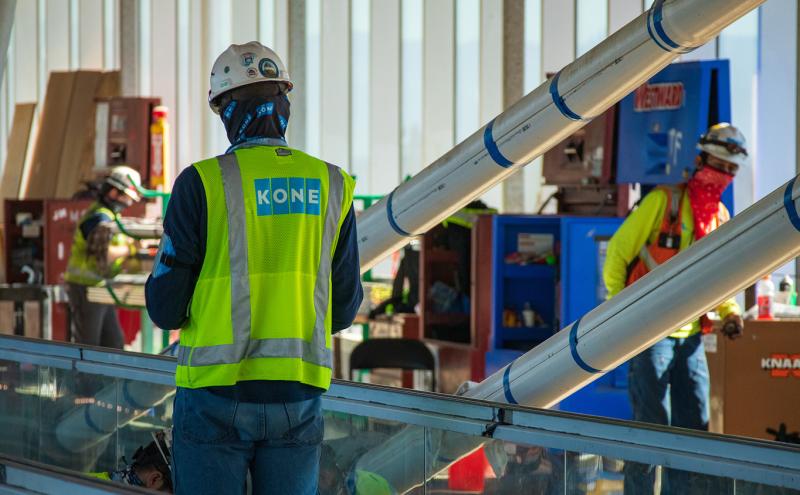 Construction workers wearing safety vests and helmets work inside a large building with metal beams, equipment cabinets, and tools visible in the background.