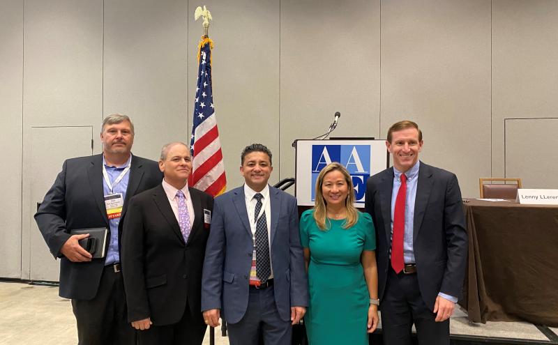 A group of five people stands together in a conference room in front of a podium displaying the AAEE logo and a U.S. flag. Name badges, conference materials, and a table with a nameplate are visible in the background.