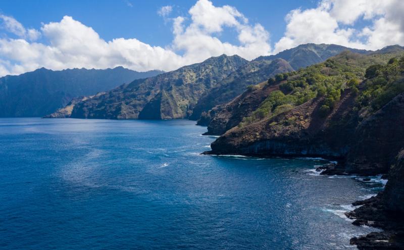 Coastal cliffs rising above deep blue ocean under a bright sky with scattered clouds.
