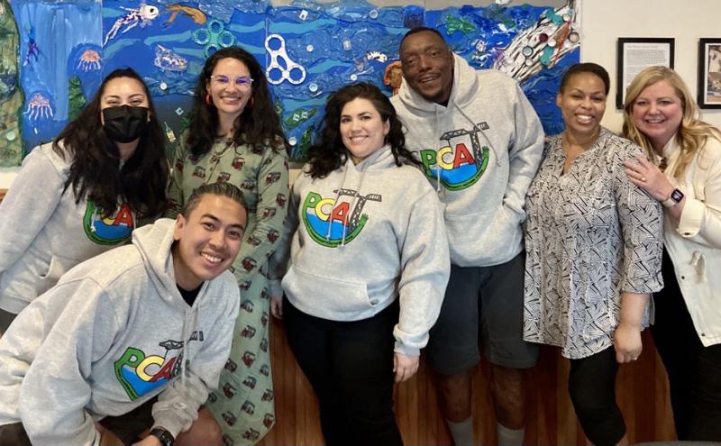 A group of people standing together indoors in front of a colorful wall display, with several wearing hoodies featuring the letters “PCA.