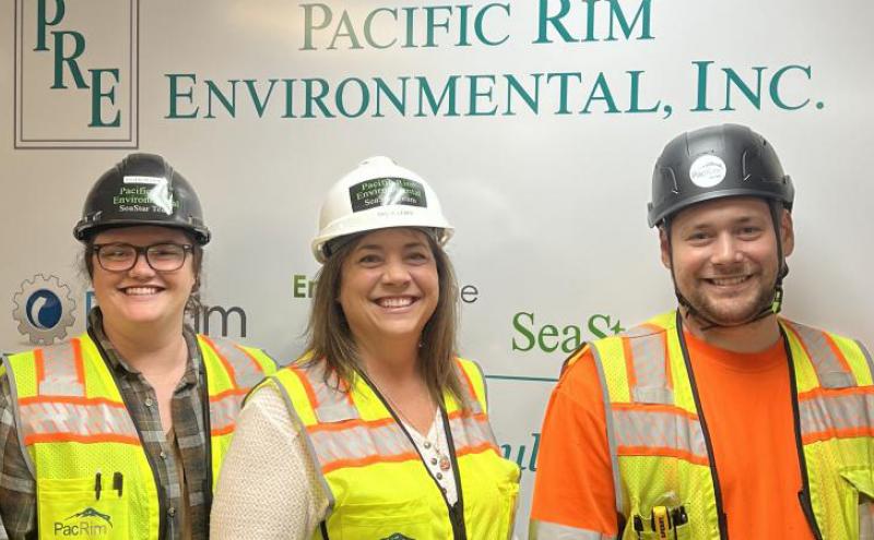 Three people wearing safety vests and hard hats standing in front of a sign for “Pacific Rim Environmental, Inc.