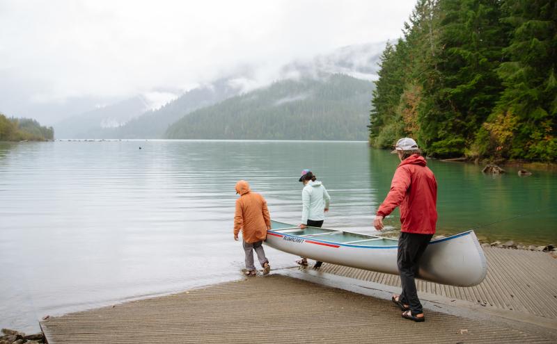 Three people wearing outdoor jackets carry a canoe down a boat ramp toward a calm, misty lake surrounded by forested mountains.