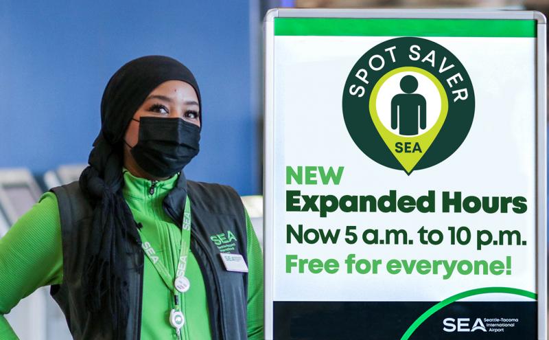 An SEA Airport employee standing next to a sign reading 'Spot Saver SEA: New Expanded Hours - Now 5 a.m. to 10 p.m. Free for everyone!'