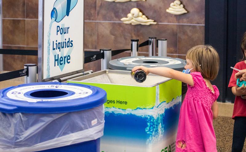 Child pouring liquid from a bottle into a designated liquids disposal station at an airport, next to recycling and trash bins.