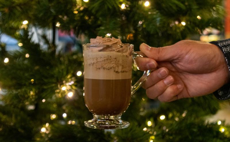 Hand holding a glass mug of a whipped‑topped coffee drink in front of a lit holiday tree.