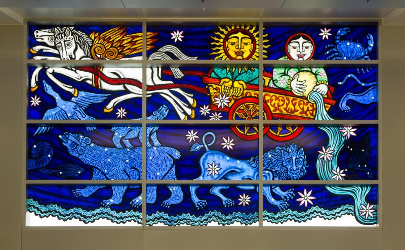 A section of stained glass showing winged horses pulling a chariot carrying the sun and the moon, with constellation animals below. 