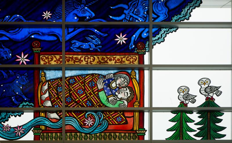 Stained glass depicting two figures sleeping in an decorated bed, with two owls perched on an evergreen tree besides them.