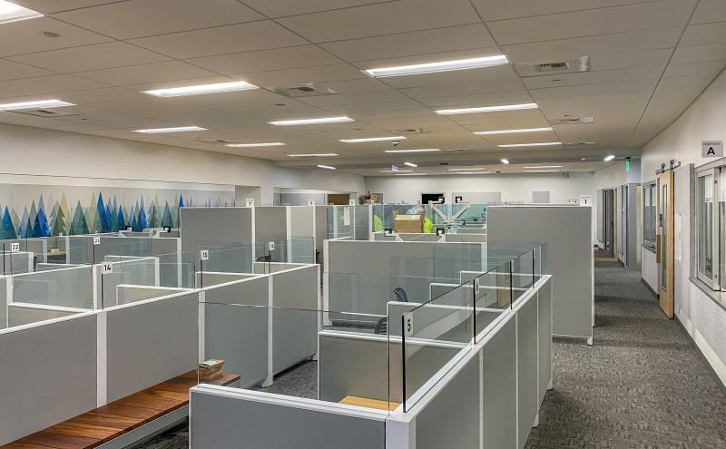 A large indoor office space with rows of light gray cubicles separated by glass panels, each labeled with numbers. The area is well-lit with ceiling lights, and a hallway runs along the right side of the room. A wall on the left features a decorative mural with blue and green triangular shapes.