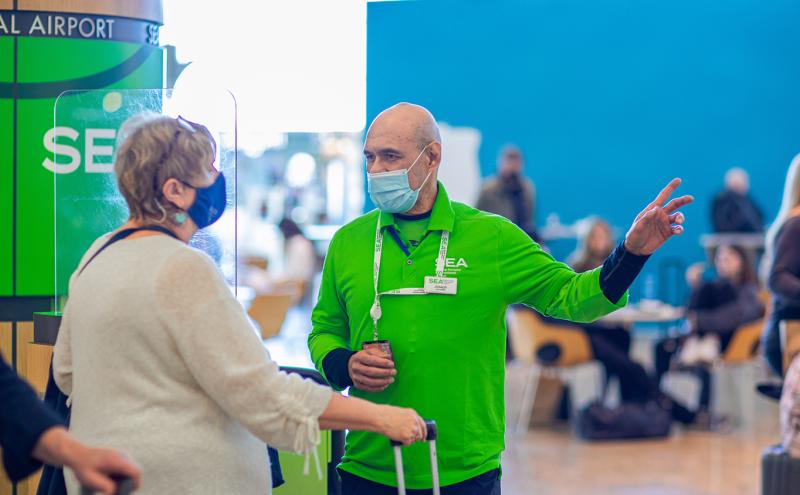 A person wearing a bright green shirt and a face mask is speaking with another person in an airport area, with signs, seating, and travelers visible in the background.