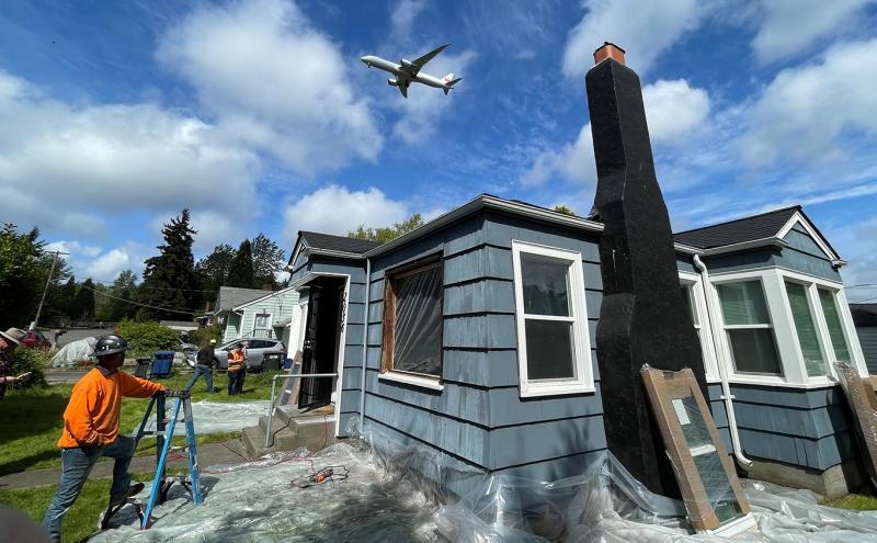 Workers outside a small house undergoing renovation, with plastic sheeting around the foundation and ladders and tools on the lawn, while an airplane flies overhead in a partly cloudy sky.
