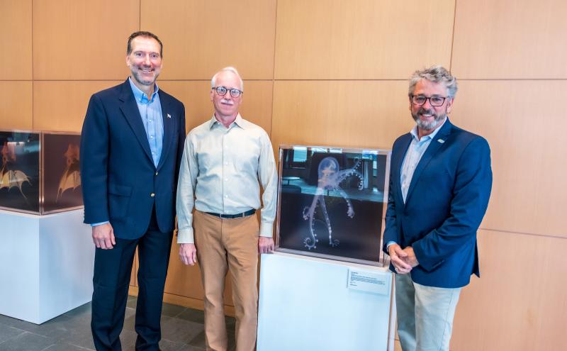 Alt text: Three individuals stand in a lobby area beside display cases showing large photographic images of deep‑sea organisms against a light wood wall.