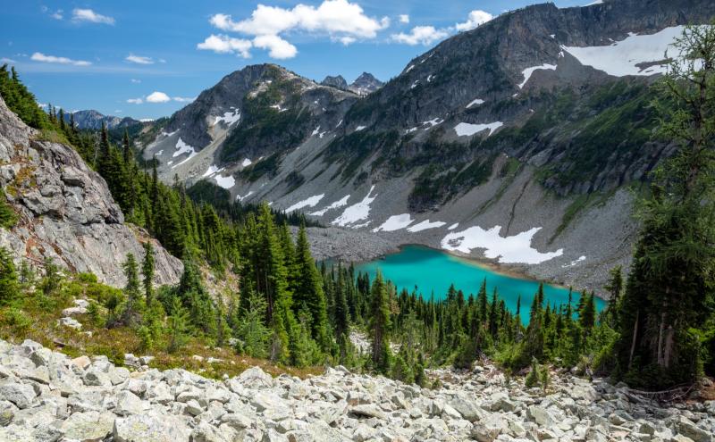 Mountain landscape with a turquoise alpine lake surrounded by rocky slopes, evergreen trees, and patches of snow under a blue sky.