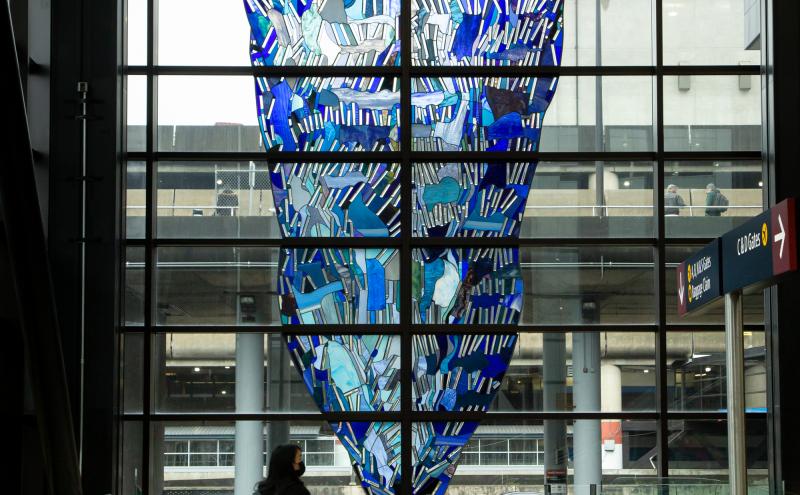 For A.W. A stained-glass window in an inverted triangle form, installed in a floor-to-ceiling airport window wall, with a traveler visible in silhouette at the base.  