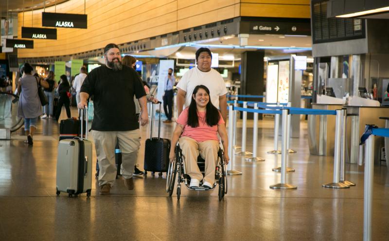 “Interior airport scene showing a person using a wheelchair being assisted by two people, all moving through a terminal area with luggage. Overhead signs display airline information, and stanchions form queue lines in the background.