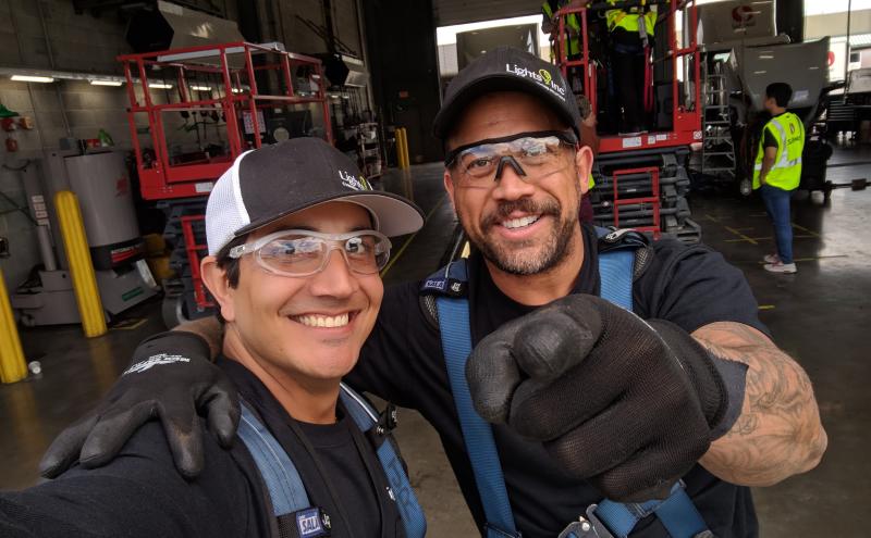 Two workers wearing safety gear take a selfie inside a warehouse, with lift equipment and additional workers visible in the background.