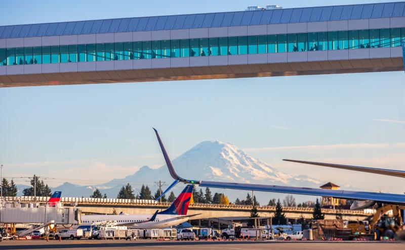 An airport scene with airplanes on the tarmac and a pedestrian skybridge above, with a snow-covered mountain visible in the background.