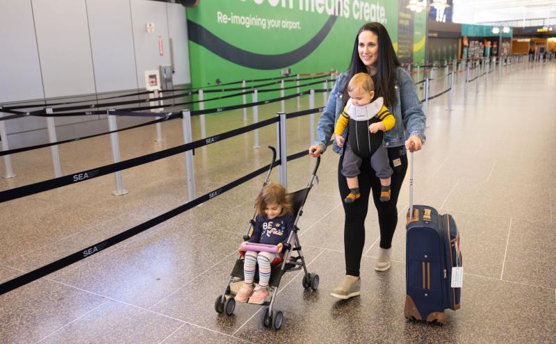 A mom walks in the terminal pushing a suitcase and a toddler in an umbrella stroller while also wearing a baby in a carrier. 